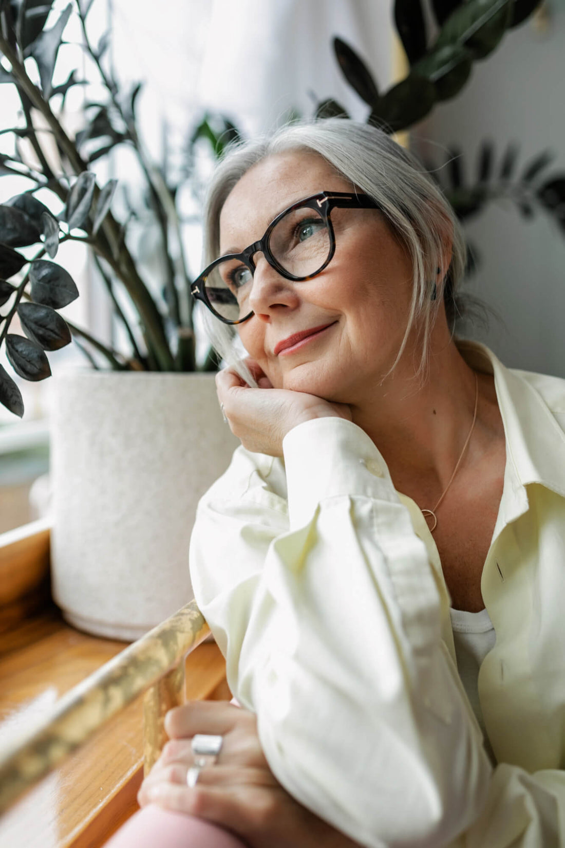Woman with glasses leaning on a surface with plants in the background